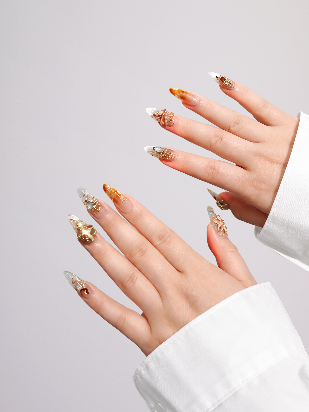 A pair of hands with long, sculpted almond-shaped nails decorated with gold glitter, metallic patterns, and clear tips, styled with a white long-sleeve shirt against a light gray background.