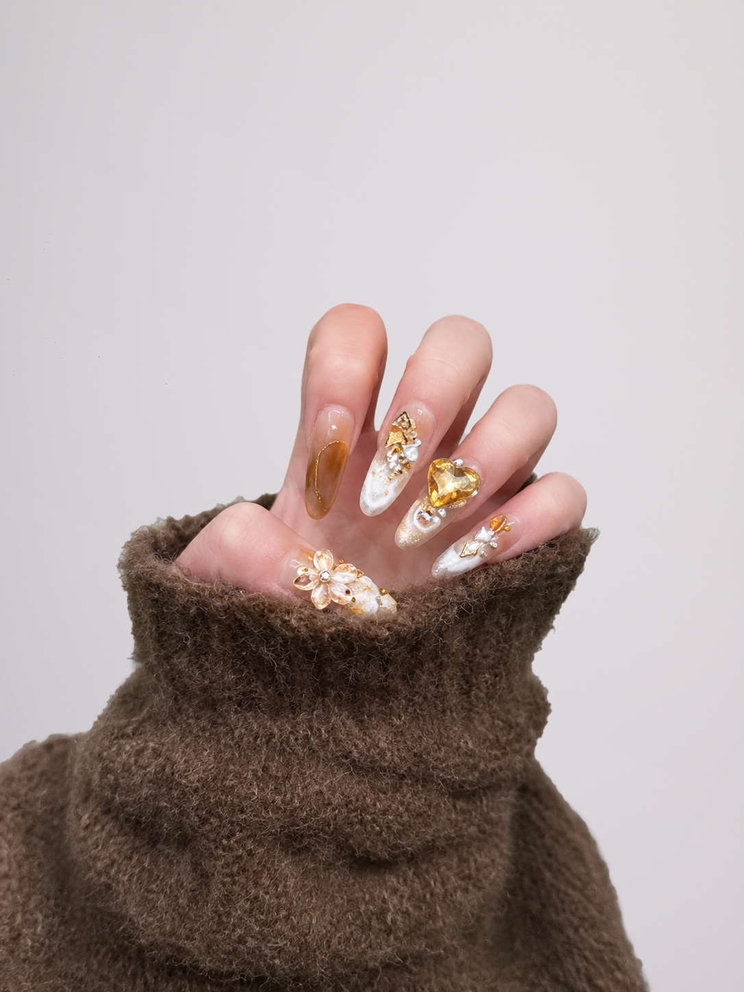 A close-up of a hand with long, decorated press-on nails, wearing a brown fuzzy knit sleeve, set against a plain light gray background.