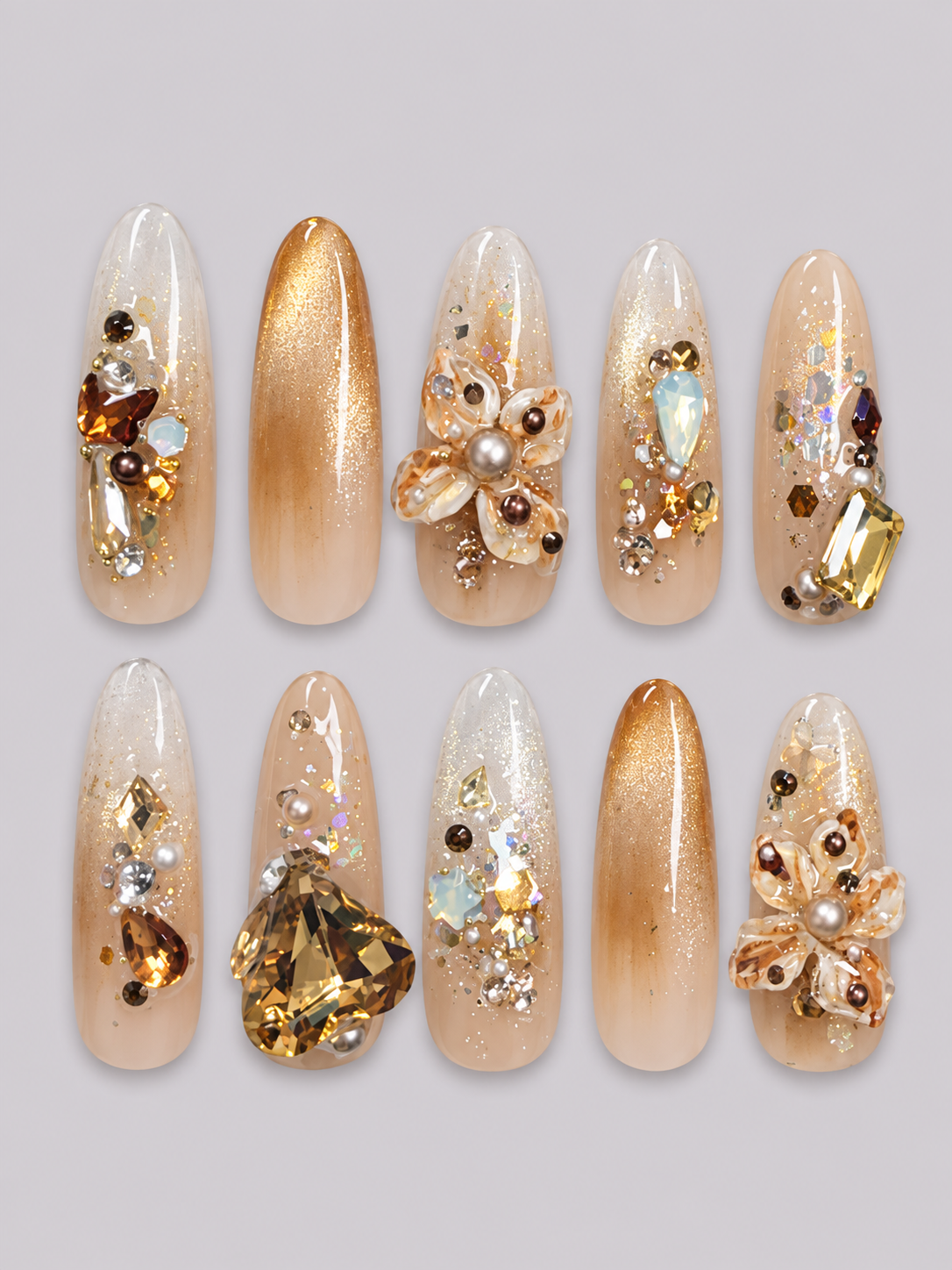A pair of hands with long, sculpted almond-shaped nails decorated with gold glitter, metallic patterns, and clear tips, styled with a white long-sleeve shirt against a light gray background.