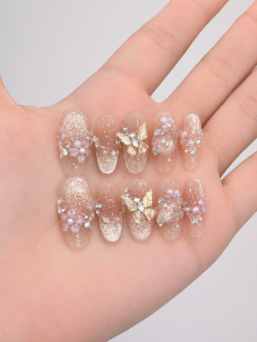A hand displaying a set of decorative press-on nails. The nails are clear and transparent with a glittery gold base, decorated with small pink pearls, clear rhinestones, and gold butterfly embellishments.