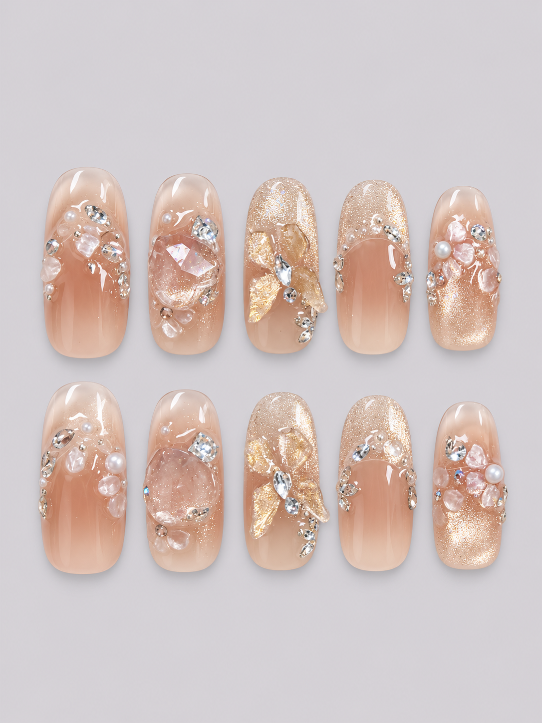 A hand displaying a set of decorative press-on nails. The nails are clear and transparent with a glittery gold base, decorated with small pink pearls, clear rhinestones, and gold butterfly embellishments.
