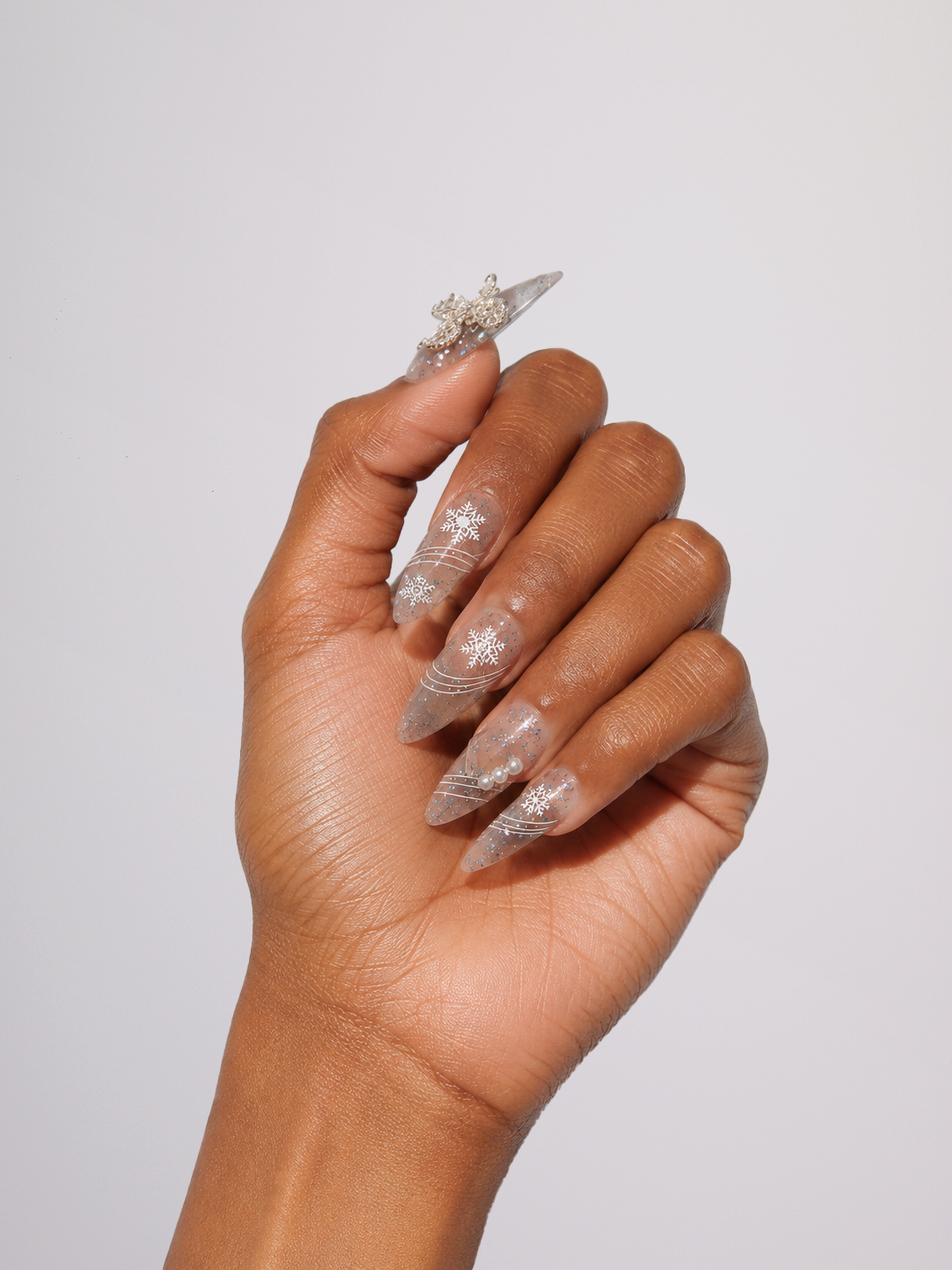 A hand with long, sculpted clear acrylic nails decorated with intricate silver glitter and crystal embellishments, shown against a plain light gray background.