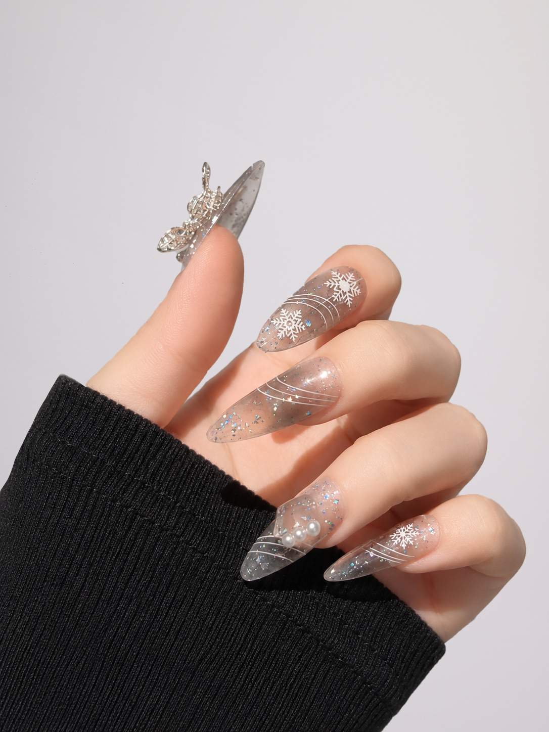 A close-up of a hand wearing long, clear stiletto-style press-on nails with silver glitter and swirl patterns, along with a silver ring, against a light gray background.