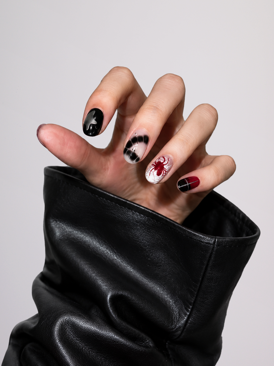 A close-up of a hand with gothic-themed nail art, featuring black, white, and red designs, wearing a black faux-leather sleeve against a light gray background.