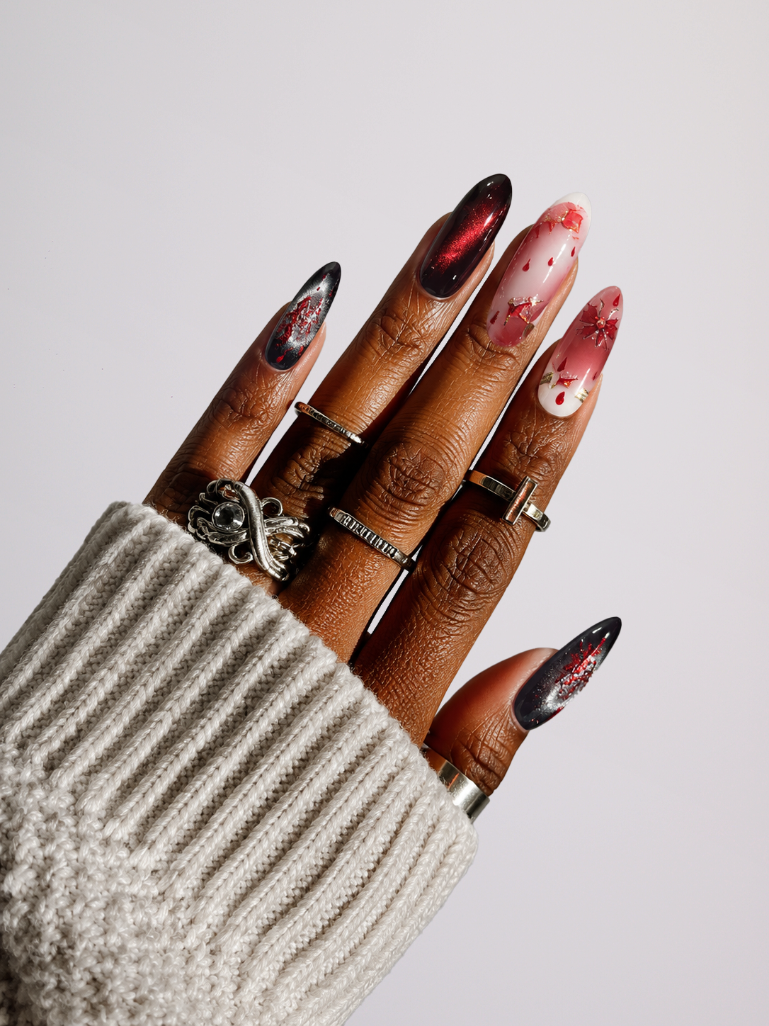 A close-up of a hand wearing a beige ribbed knit sweater sleeve and multiple silver rings, with long almond-shaped nails decorated in black, red, and white patterns against a light gray background.