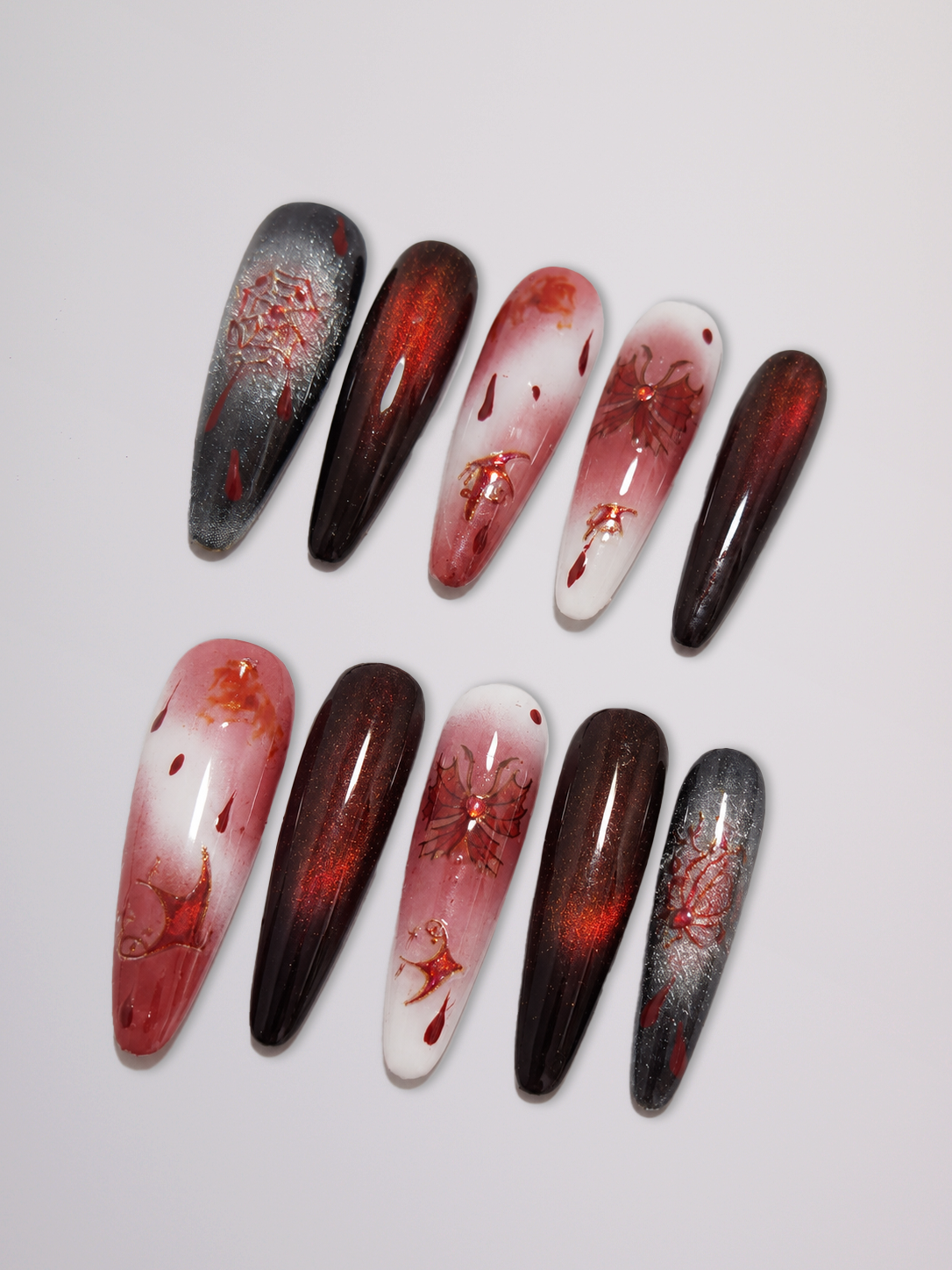 Ten long coffin-shaped press-on nails arranged in two rows on a light gray background. The nails feature various designs including solid dark brown, dark gray with red splatters, and red-to-white gradient patterns with red splatters.
