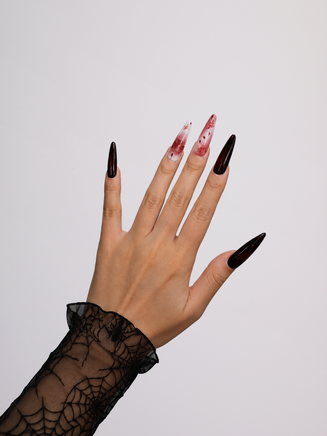 A hand wearing long, stiletto-shaped black nails with two alternating nails featuring a red and white marbled pattern, paired with a black lace cuff against a plain light gray background.