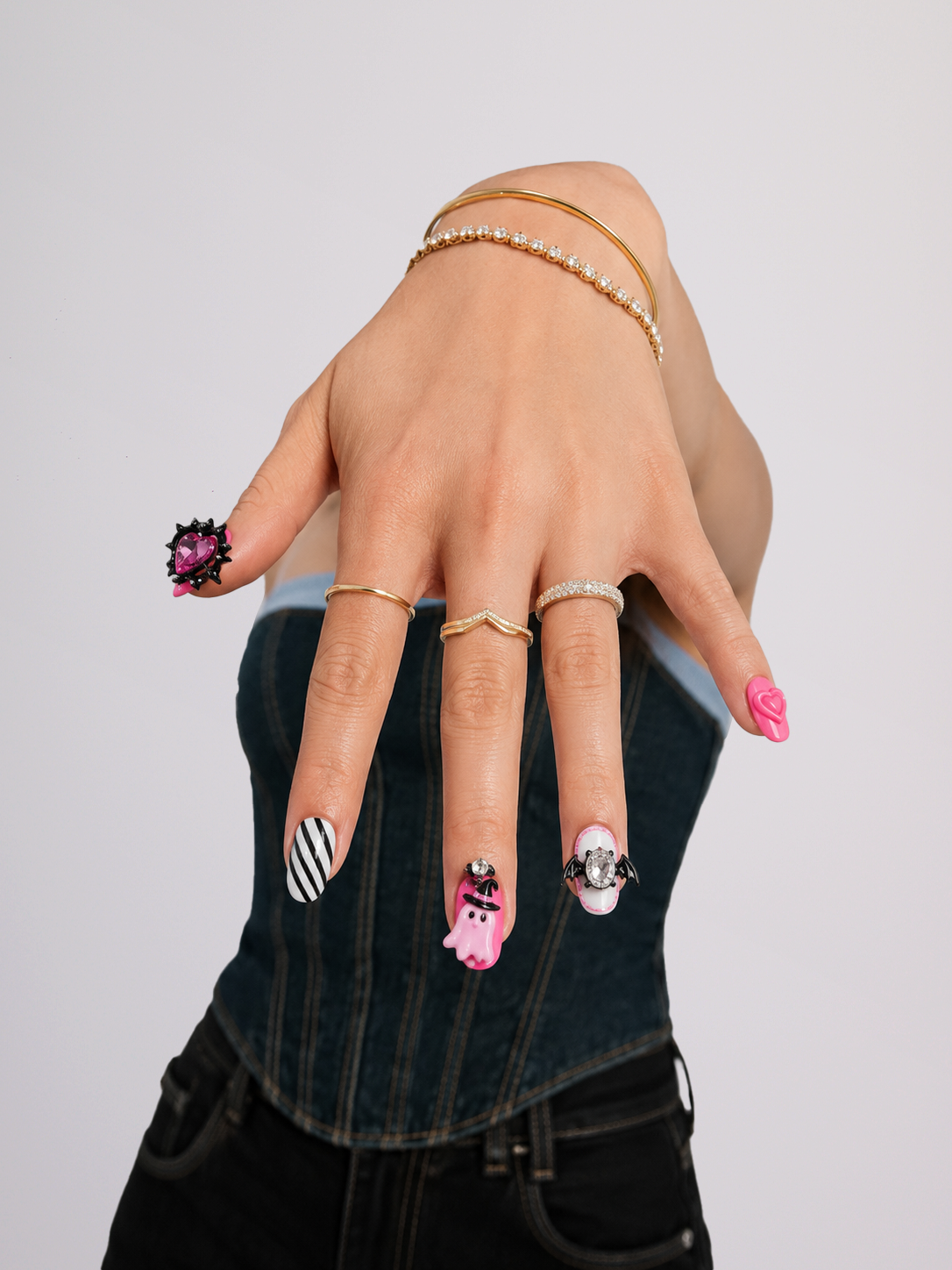 A close-up of a hand wearing multiple rings, including a large black and white striped ring, a pink ring with a skull design, and a silver ring with a skull and crossbones, against a light gray background.