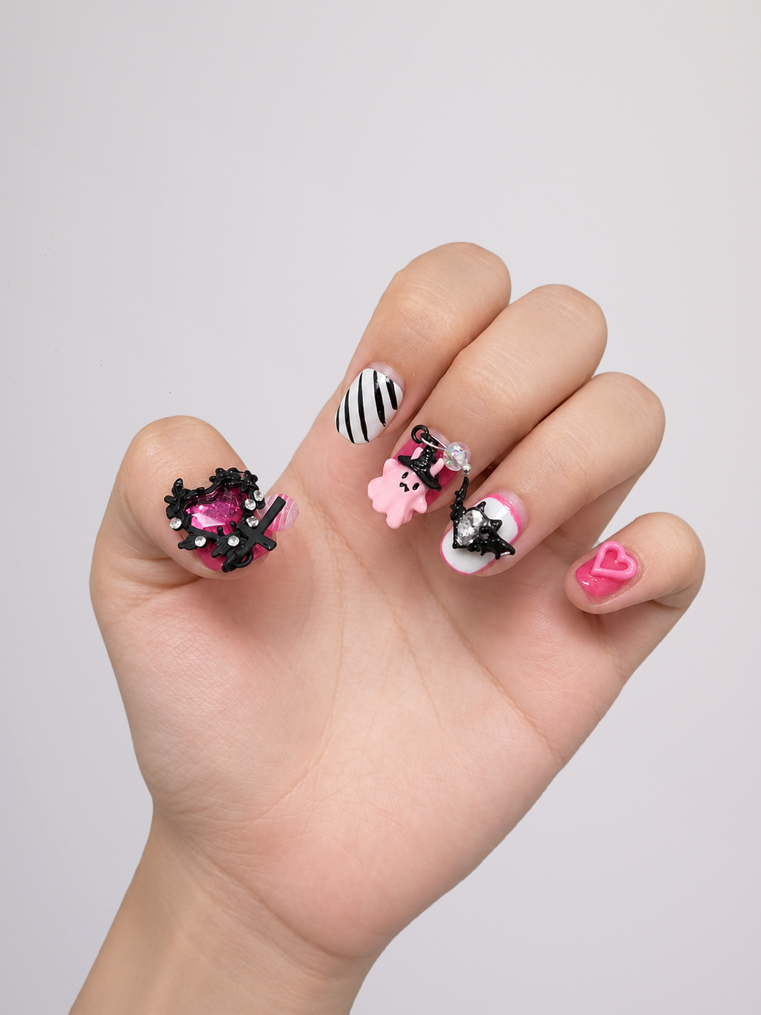 A hand displaying a set of decorative, gothic-themed press-on nails against a plain light gray background. The nails feature black lace, pink gems, black-and-white stripes, a pink cartoon character, and a pink heart design.