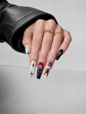 A close-up of a hand with long coffin-shaped nails featuring gothic designs, including black crosses on white and black nails, and a red, white, and blue splatter pattern on one nail. The hand is wearing gold rings and a black leather sleeve, set against a neutral gray background.