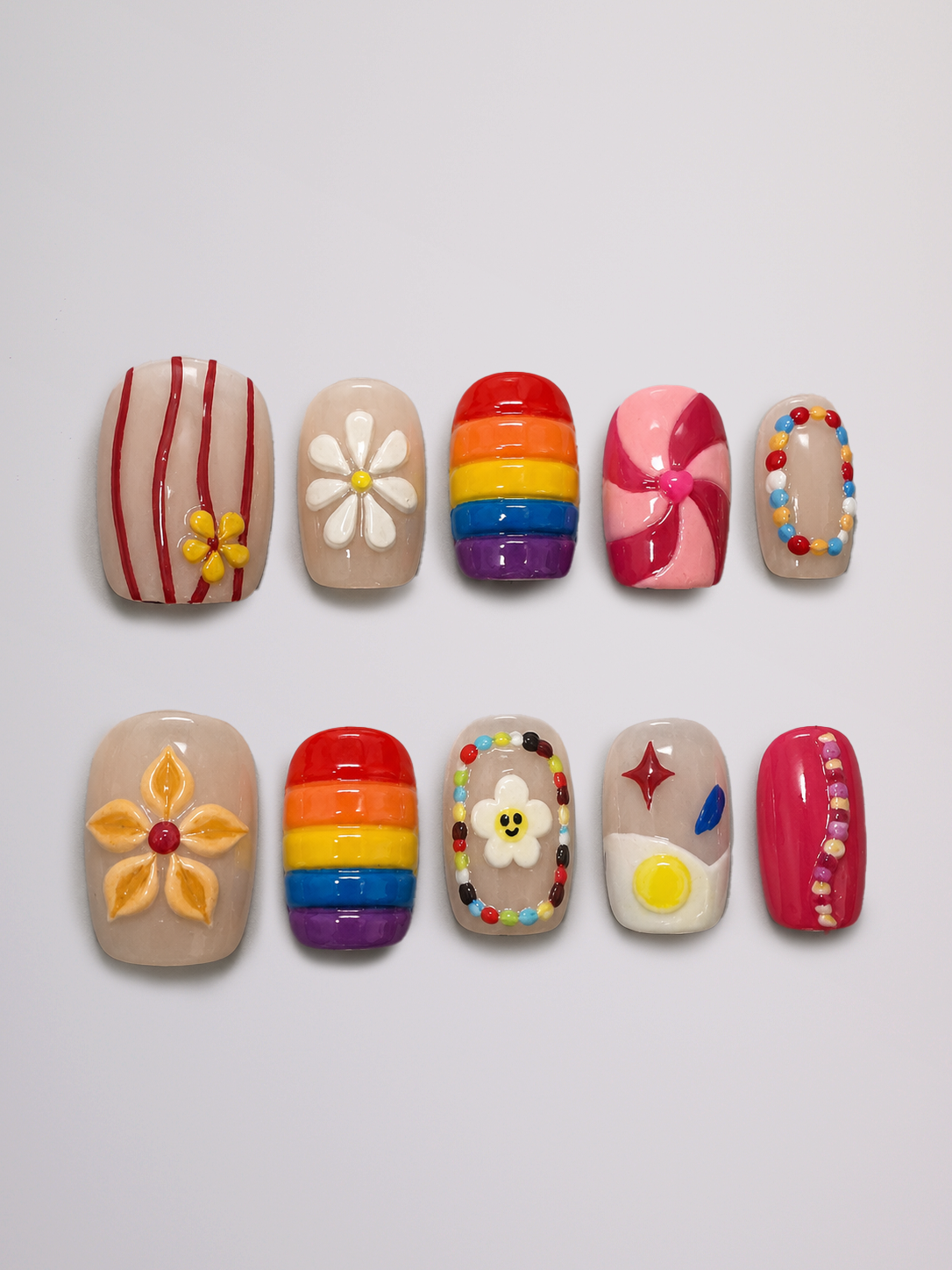 Ten artificial press-on nails arranged in two rows on a light gray background. The nails feature various colorful designs including stripes, flowers, rainbow patterns, swirls, and dots.