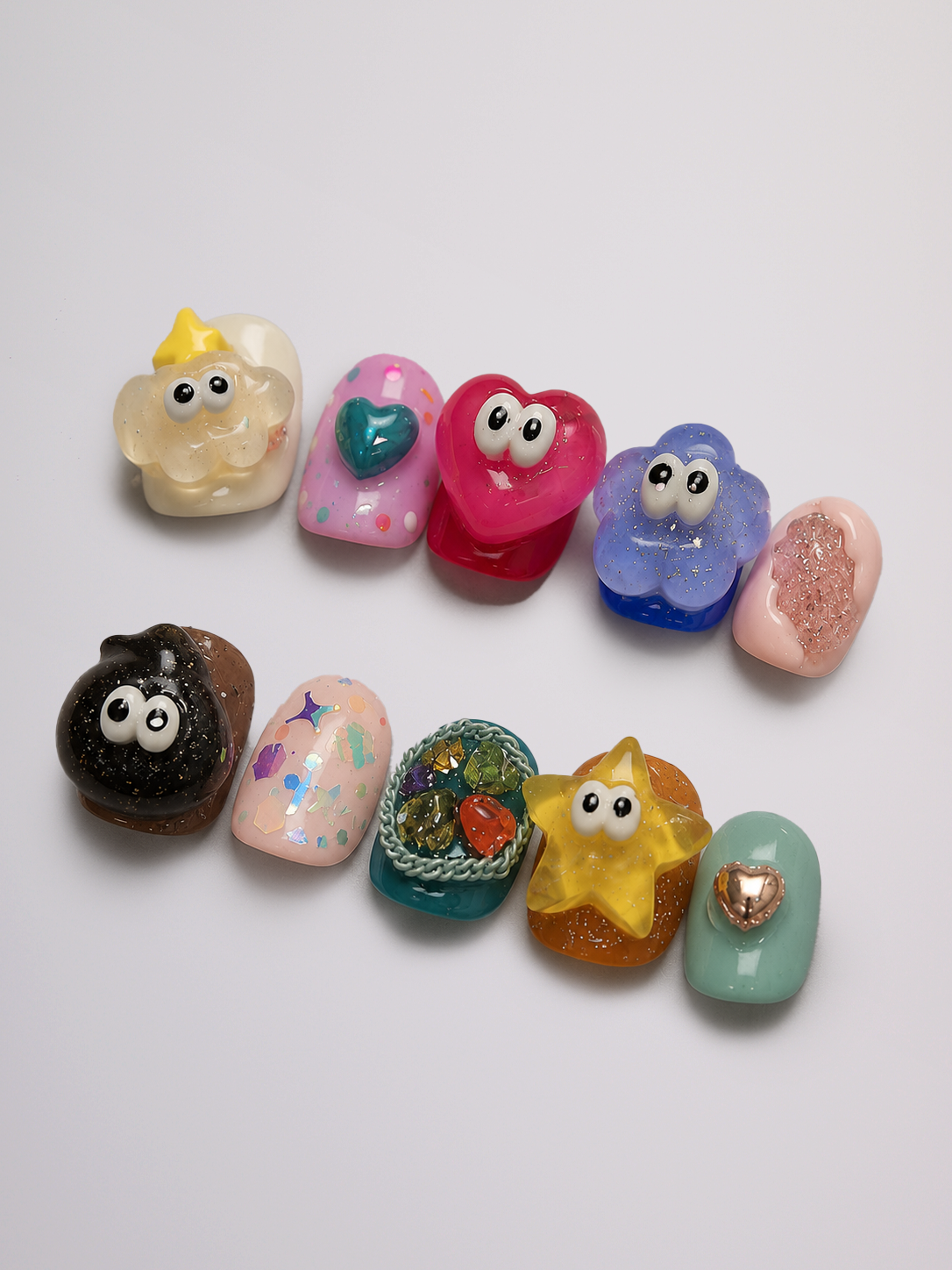 Ten colorful, cartoon-style false nail tips arranged in two rows on a plain white background. The designs include a cream cloud with a yellow crown, pink and red hearts with eyes, blue and purple flowers with eyes, a black cloud with eyes, a pink speckled heart, a green basket with colorful items, a yellow star with eyes, a mint green oval with a heart, and a pink cloud with a gold heart.