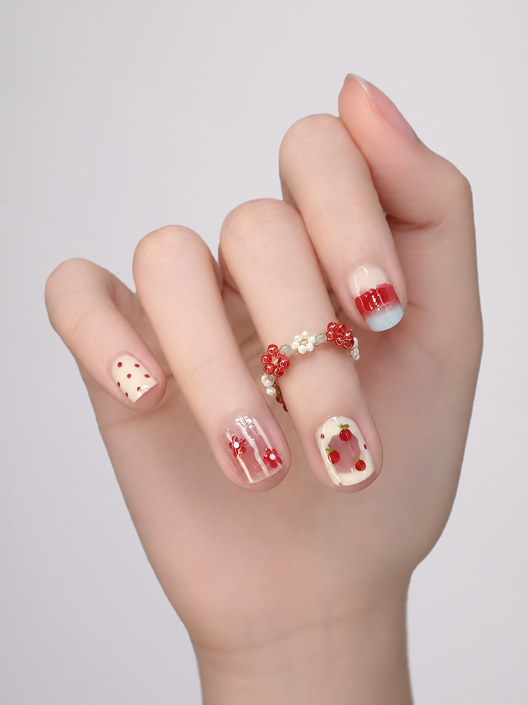 A close-up of a hand showcasing a variety of nail art designs, including strawberry patterns, red stripes, and small red hearts, with a red and white beaded ring on one finger, set against a plain light gray background.