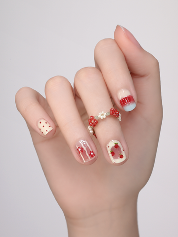 A close-up of a hand showcasing a variety of nail art designs, including strawberry patterns, red stripes, and small red hearts, with a red and white beaded ring on one finger, set against a plain light gray background.