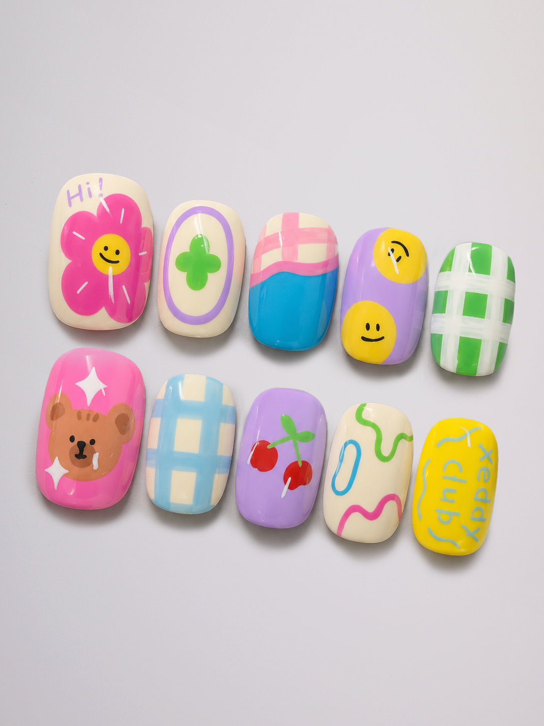 A set of ten decorative false nails arranged in two rows on a light gray background. The nails feature pastel colors and cute designs including a pink flower with a smiley face, a green clover, pink and blue stripes, yellow smiley faces, a green and white gingham pattern, a brown bear face, red cherries, abstract squiggly lines, and yellow text reading 'Happy Sunday'.