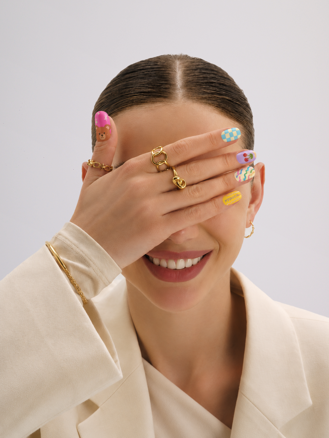 A close-up lifestyle shot of a model wearing multiple gold rings on her fingers, with her hand covering part of her face. She is wearing a cream-colored blazer and has colorful patterned nail art against a light gray background.