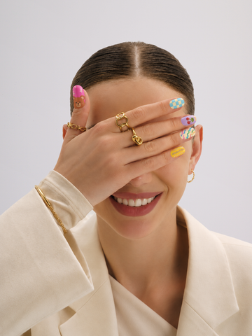 A close-up lifestyle shot of a model wearing multiple gold rings on her fingers, with her hand covering part of her face. She is wearing a cream-colored blazer and has colorful patterned nail art against a light gray background.