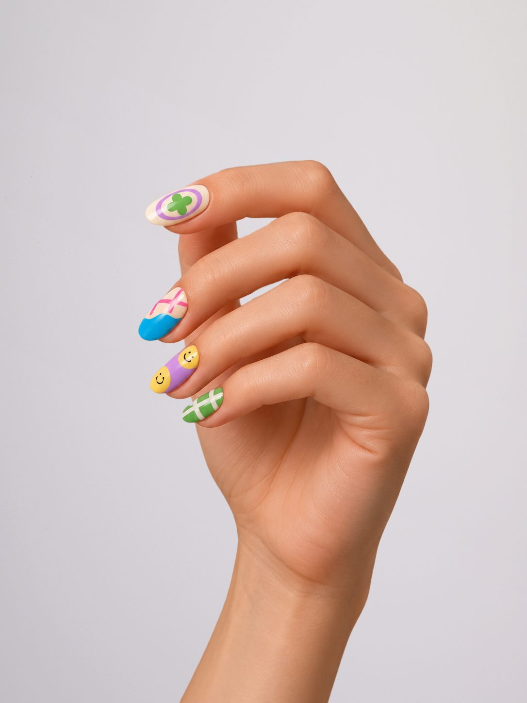 A hand with colorful Easter-themed nail art, featuring pastel designs like a green clover, pink and blue striped egg, yellow smiley face, and green-and-white stripes, set against a plain light gray background.