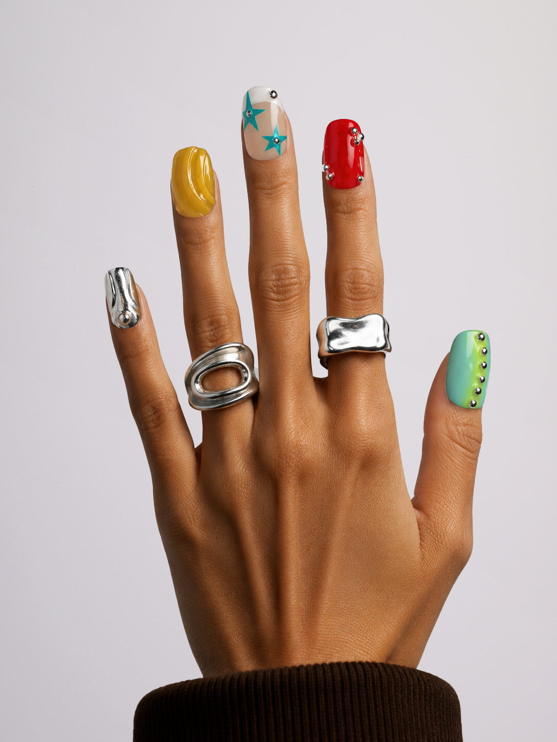 A hand with colorful manicured nails wearing multiple chunky silver rings, shown against a plain light gray background.