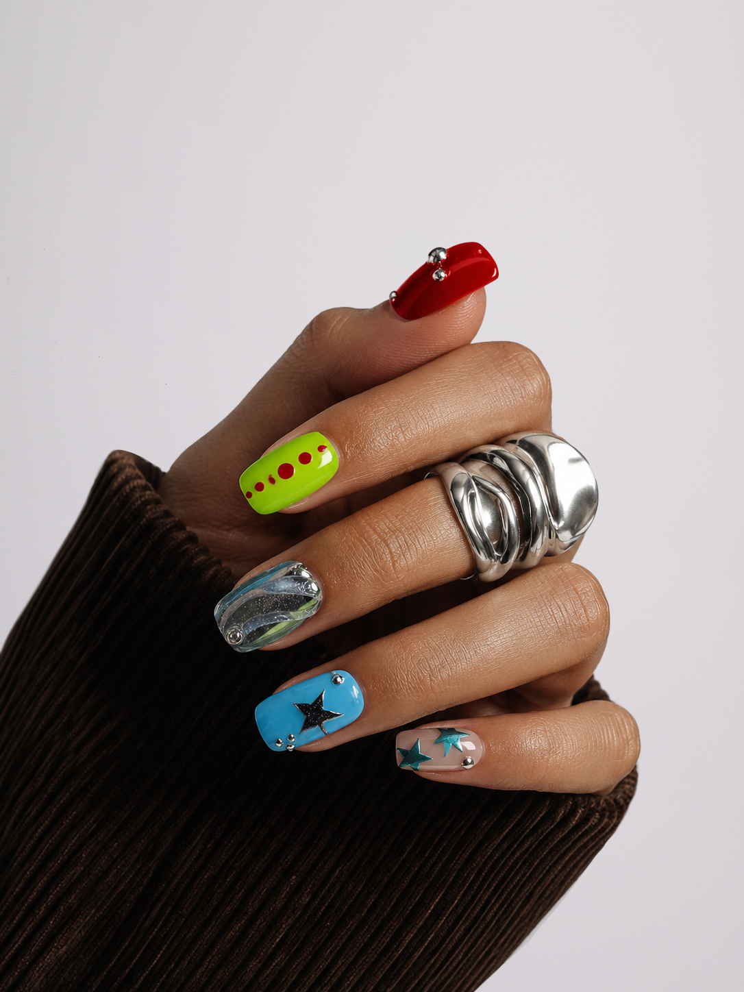 A close-up of a hand wearing a chunky silver ring and colorful nail art, including a red nail with rhinestones, a neon yellow nail with red dots, a metallic silver nail, a blue nail with a black star, and a clear glittery nail with a star.