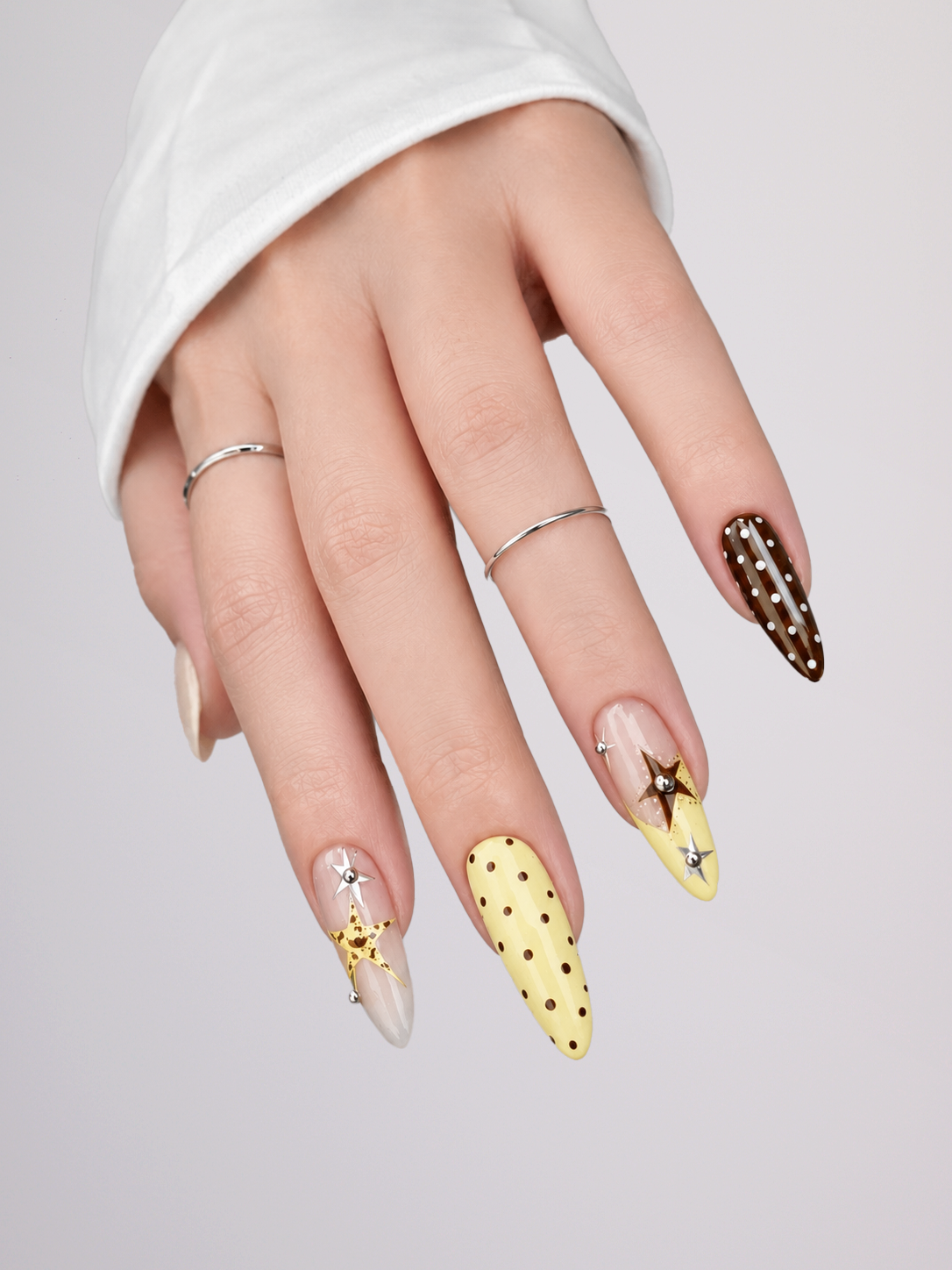 A close-up of a hand wearing long almond-shaped nails with decorative designs: one pale yellow nail with black polka dots, one brown nail with white polka dots and a star motif, and two clear/nude nails with small silver embellishments. The hand is posed against a light gray background, wearing a white ribbed sleeve.