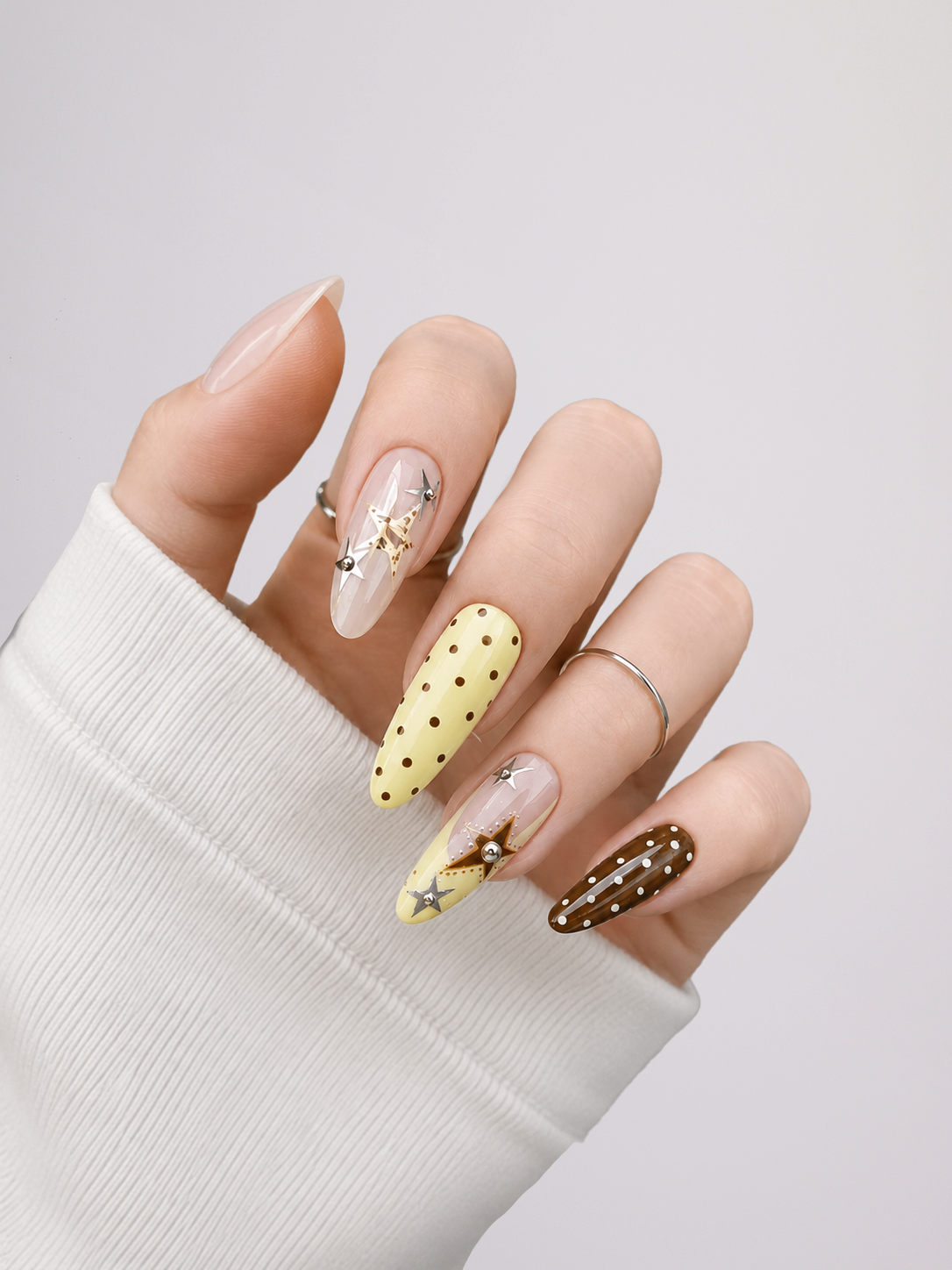 A close-up of a hand wearing long almond-shaped nails with decorative designs: one pale yellow nail with black polka dots, one brown nail with white polka dots and a star motif, and two clear/nude nails with small silver embellishments. The hand is posed against a light gray background, wearing a white ribbed sleeve.