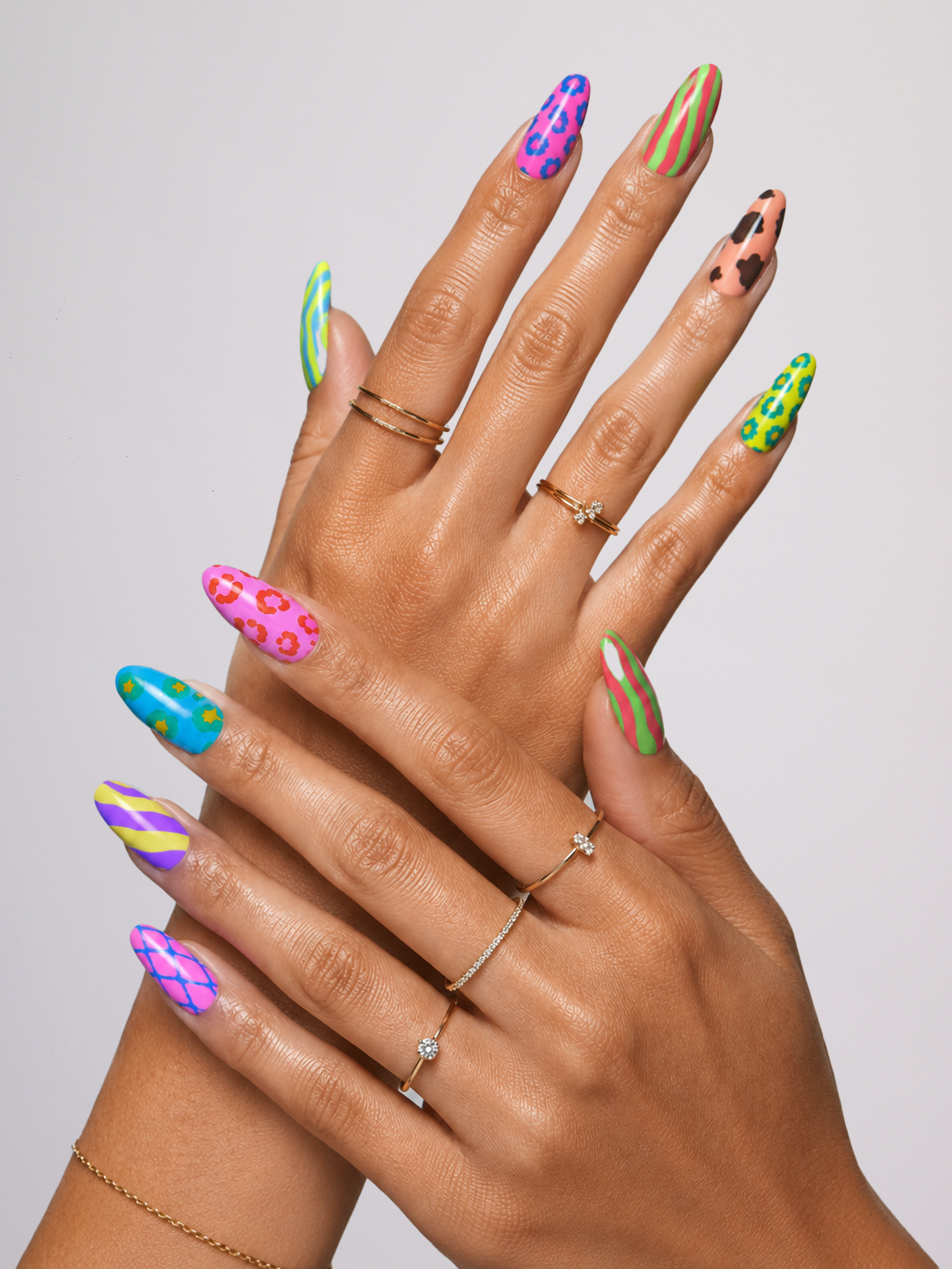 A pair of hands with long stiletto-shaped nails decorated in vibrant, multicolor patterns including pink flowers, green spots, blue and yellow stripes, and purple leopard print, styled with gold rings against a light gray background.