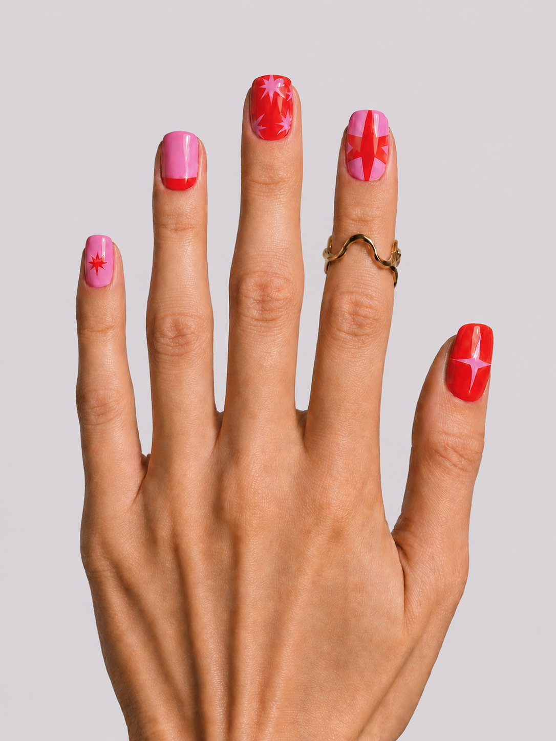 A close-up of a hand displaying a pink and red manicure with various nail art designs, wearing a gold ring against a neutral gray background.