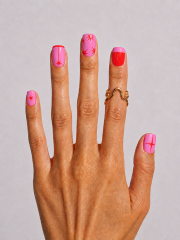 A close-up of a hand displaying a pink and red manicure with various nail art designs, wearing a gold ring against a neutral gray background.