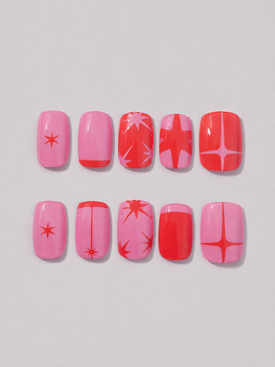 A set of ten press-on nails arranged in two rows of five on a light gray background. The nails feature various pink and red geometric patterns including stars, stripes, and starburst motifs.