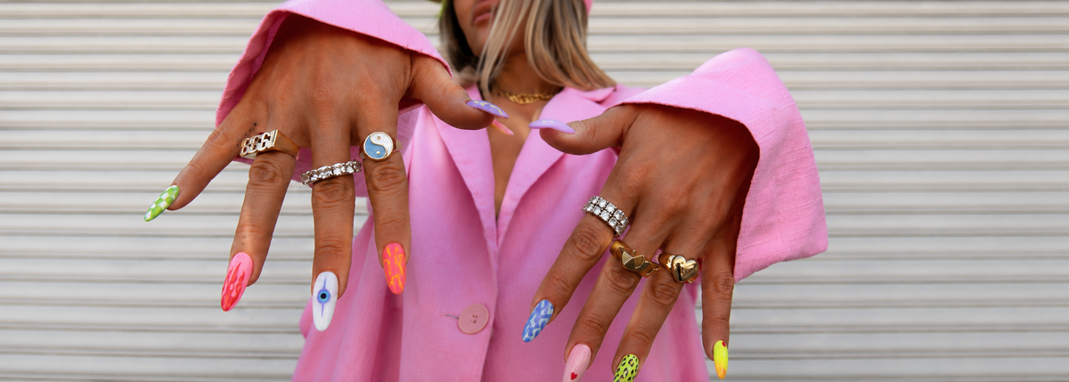 A close-up of a person wearing multiple colorful nail art designs and several silver rings, with a pink blazer against a white corrugated metal background.slider_item_NPQ49Y