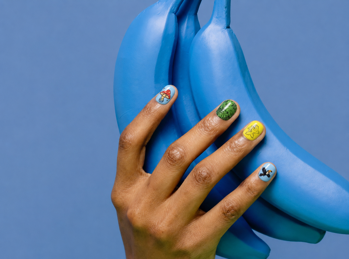 Hand wearing CLAWT x Dario Merlo press-on nails with mushroom, leaf and bird illustrations, resting on blue bananas.